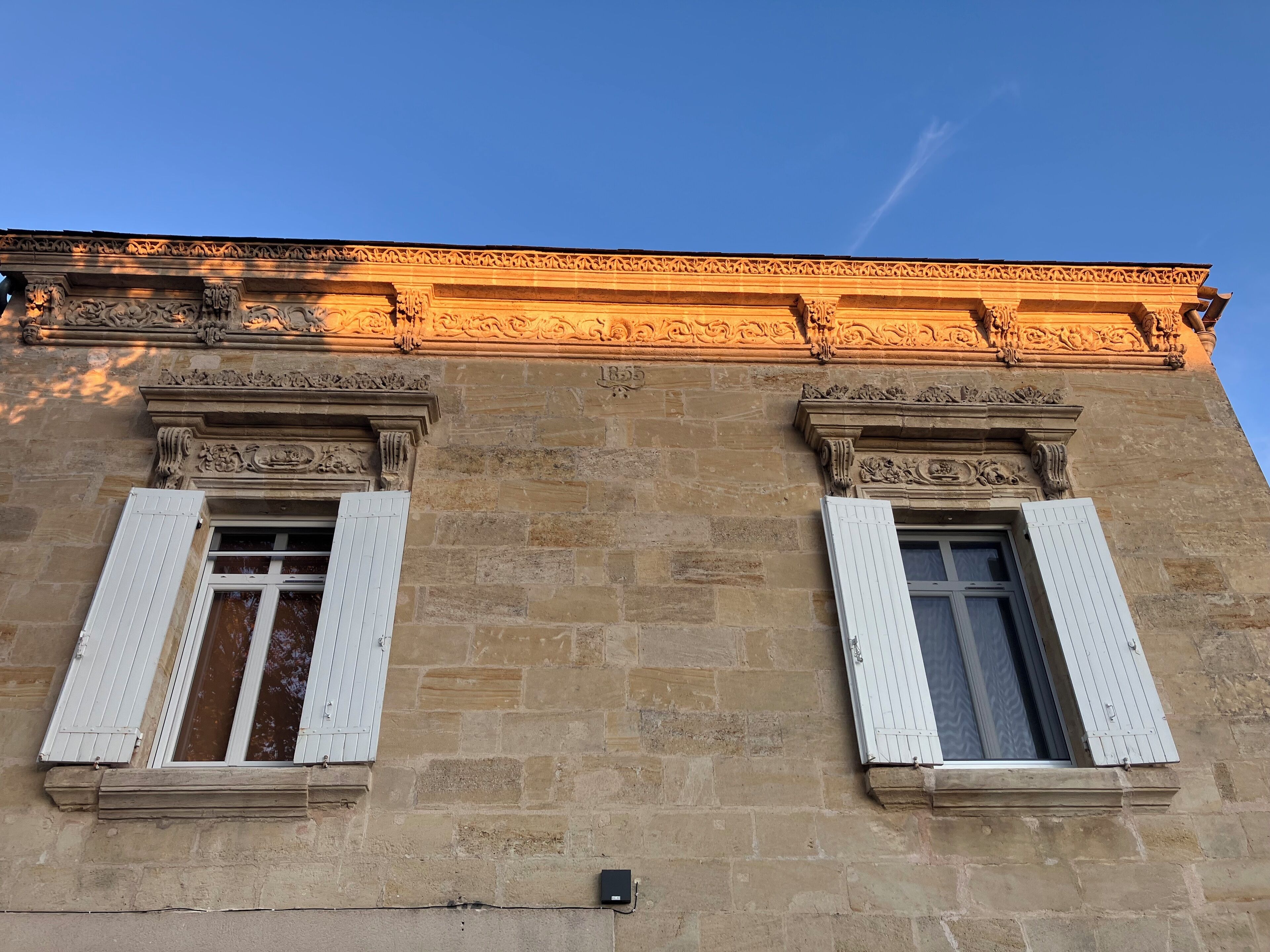 Fachada de edificio con dos ventanas y contraventanas blancas, pared de piedra y un cornisa decorativo.