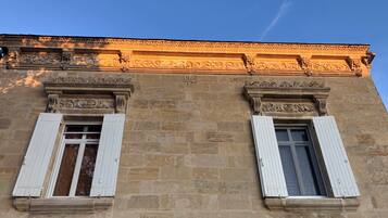 Fachada de edificio con dos ventanas y contraventanas blancas, pared de piedra y un cornisa decorativo.