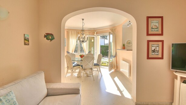 A living room with a sofa, a dining area with a table and chairs, a TV, and a hallway leading to an outdoor area. - Beautiful Villa with pool in Oliva Nova Golf (Oliva)