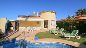 A pool area with lounge chairs, a pool ladder, and a building with a tiled roof. - Beautiful Villa with pool in Oliva Nova Golf (Oliva)