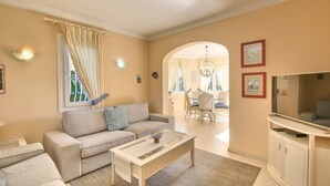 A living room with a sofa, coffee table, and TV. There is a dining area with chairs and a chandelier. - Beautiful Villa with pool in Oliva Nova Golf (Oliva)