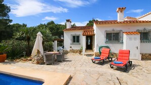 Un espace aménagé au bord de la piscine, avec des chaises longues, une terrasse en pierre et un bâtiment blanc doté d’un toit recouvert de tuiles.