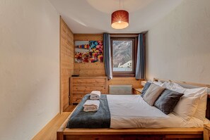 A cozy bedroom with a wooden bed, a window with a view of a mountain, and a shelf with colorful items.