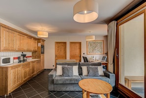 A modern kitchen with wooden cabinets, a microwave, and a coffee maker. There is a grey sofa with cushions and a small wooden table.