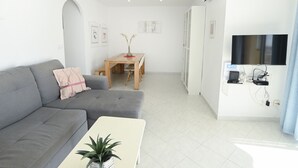 A modern living room with a grey sofa, a small white table with a plant, a wooden dining table with chairs, a TV mounted on the wall, and a white air conditioning unit. - A90 EDIFICIO IFACH III 7º 36 (Calpe / Calp)