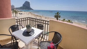 A balcony with a table and chairs, overlooking a beach with palm trees and a rocky formation in the distance. - A85 EDIFICIO OCEANIC 304 (Calpe)