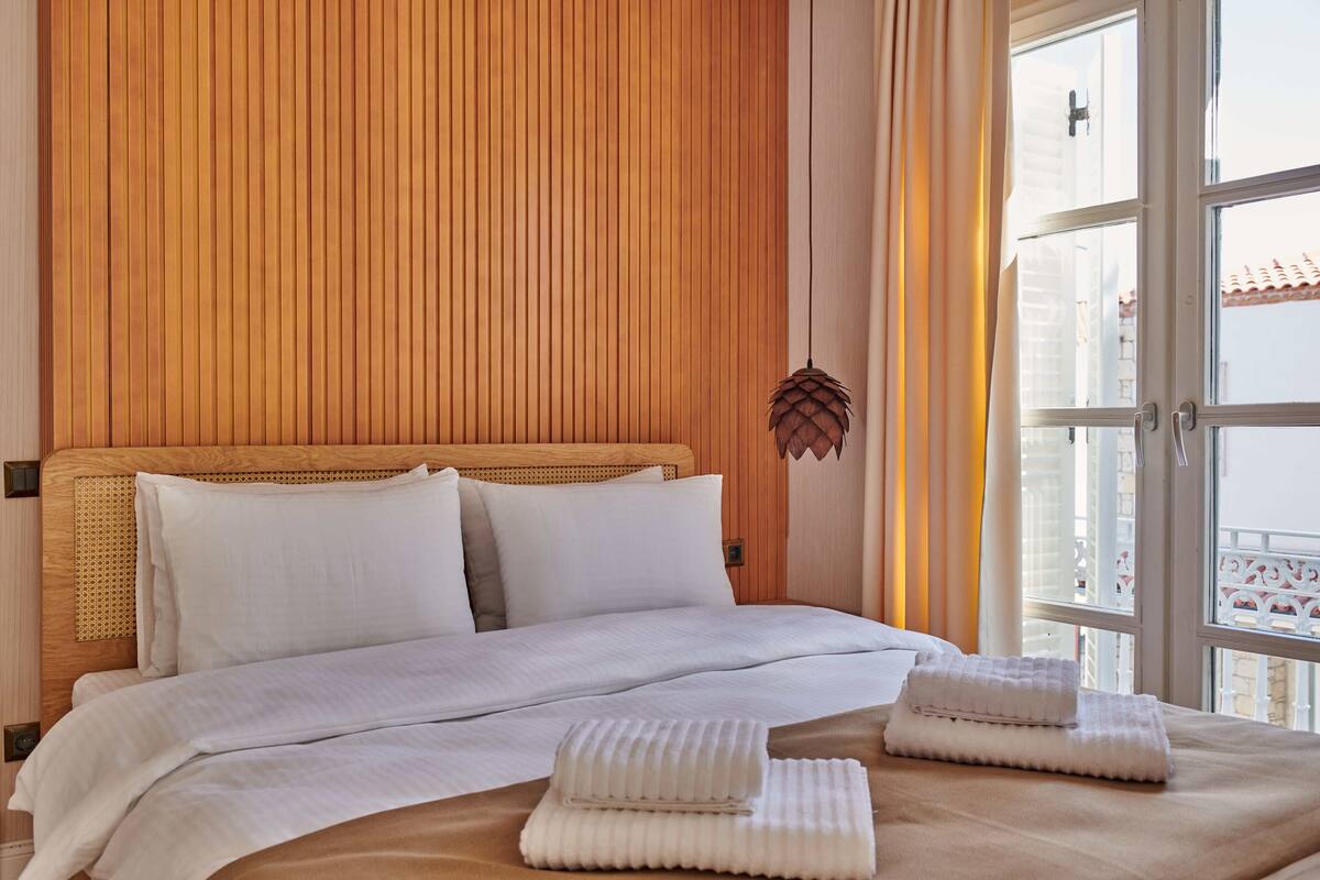 A hotel room with a wooden headboard, white bedding, and a view of a building through a window.