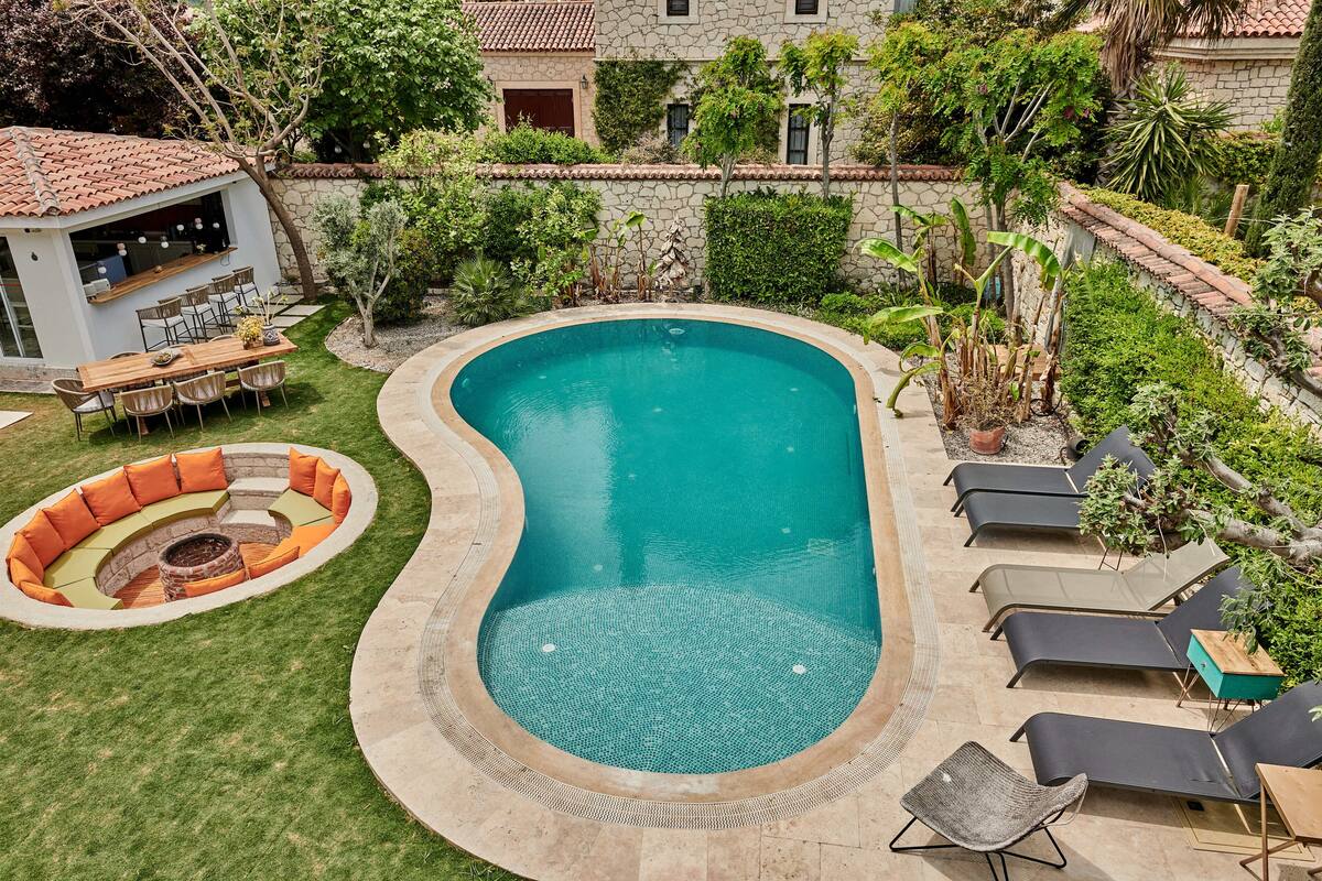 A swimming pool with a curved edge, surrounded by a grassy area and a seating area with a fire pit.
