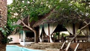 Un área de piscina con suelo de piedra, una estructura de techo de paja y mobiliario exterior bajo árboles.