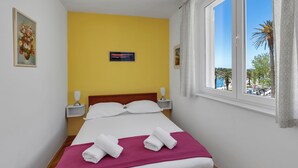 A hotel room with a bed, yellow walls, a window with a view of palm trees, and a painting on the wall. - Apartment Riva ,city center (Makarska)
