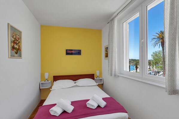 A hotel room with a bed, yellow walls, a window with a view of palm trees, and a painting on the wall. - Apartment Riva ,city center (Makarska)