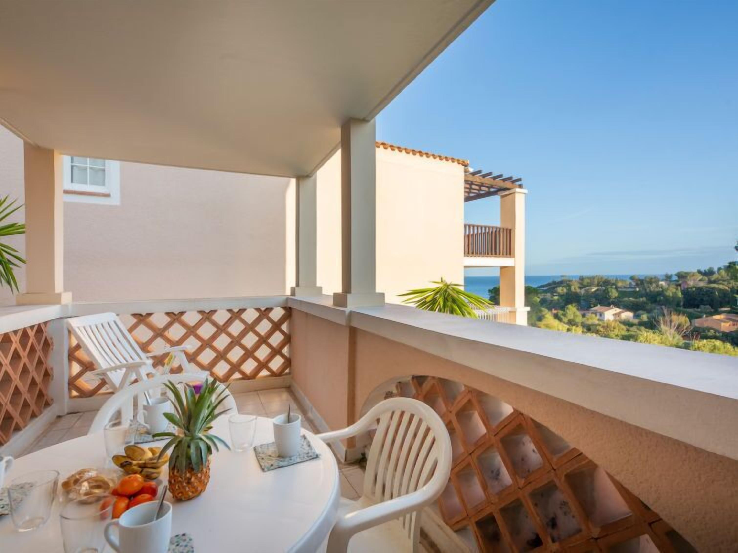 Ein Balkon mit Tisch, gedeckt für zwei Personen, mit einer Ananas, Früchten und Getränken. Der Blick umfasst eine Küstenlandschaft und ein Gebäude mit einem Ziegeldach.