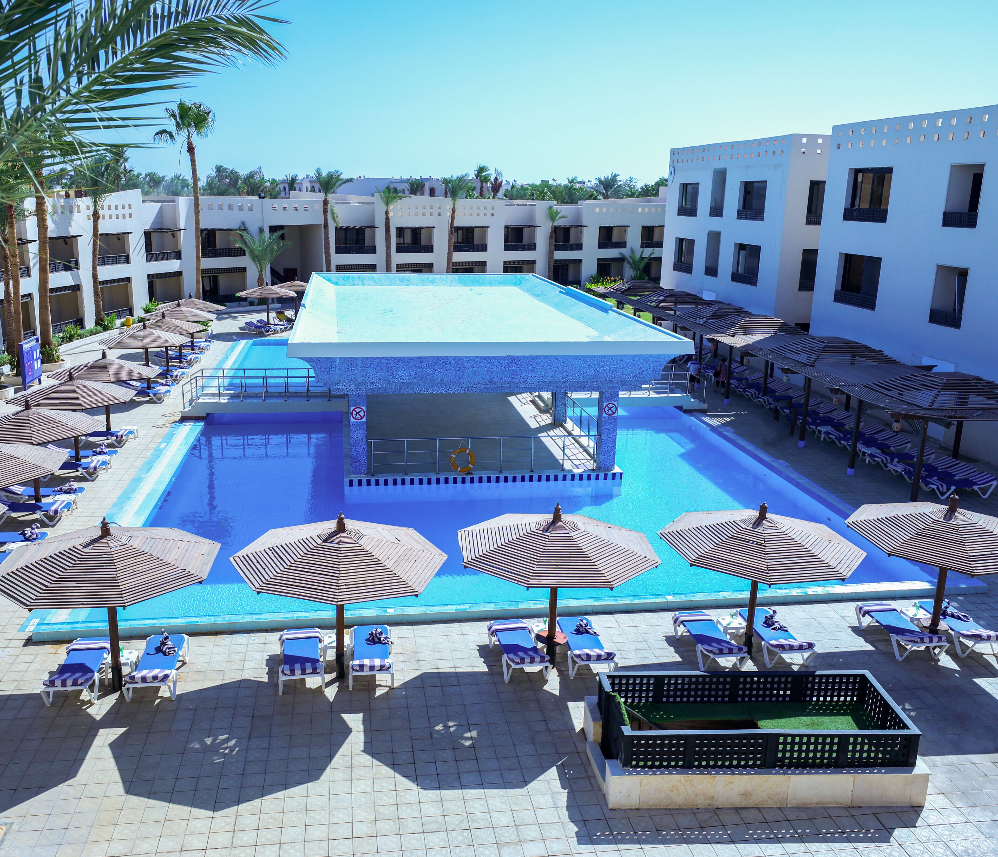 A hotel pool area with multiple lounge chairs and umbrellas, a central pool, and a building in the background.