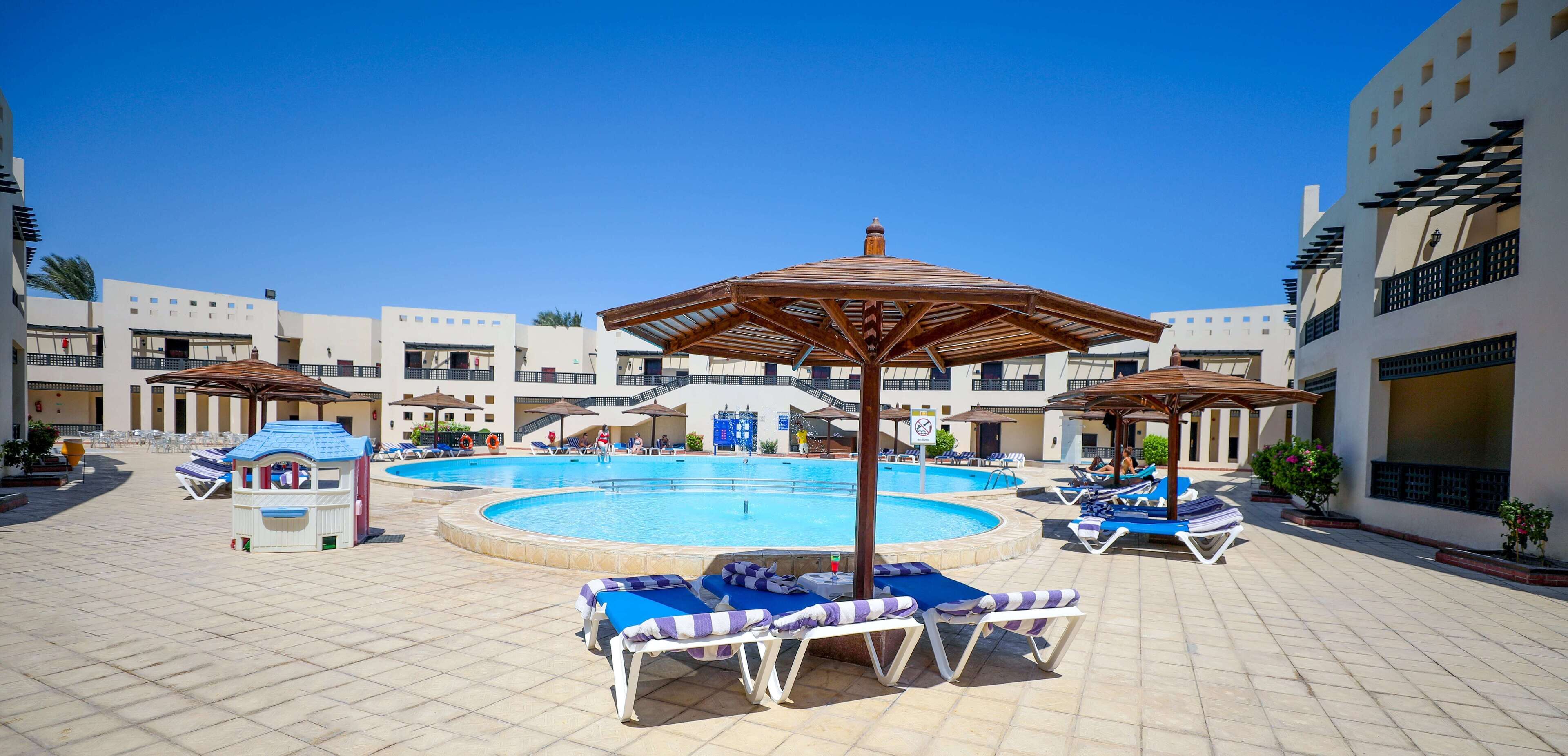 Zona de piscina amb gandules i ombrelles, envoltada d'edificis amb balcons.