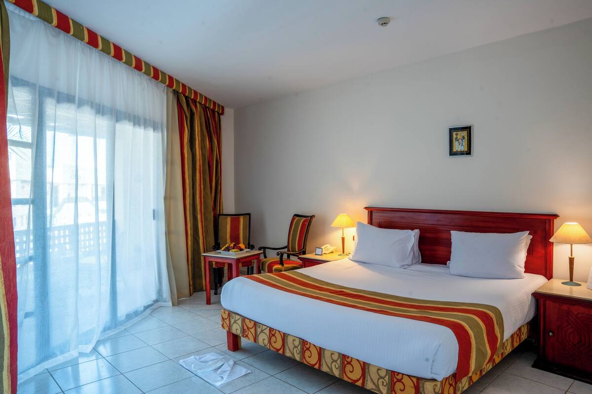 A hotel room with a large bed, a desk, a chair, a nightstand, and a window with curtains.