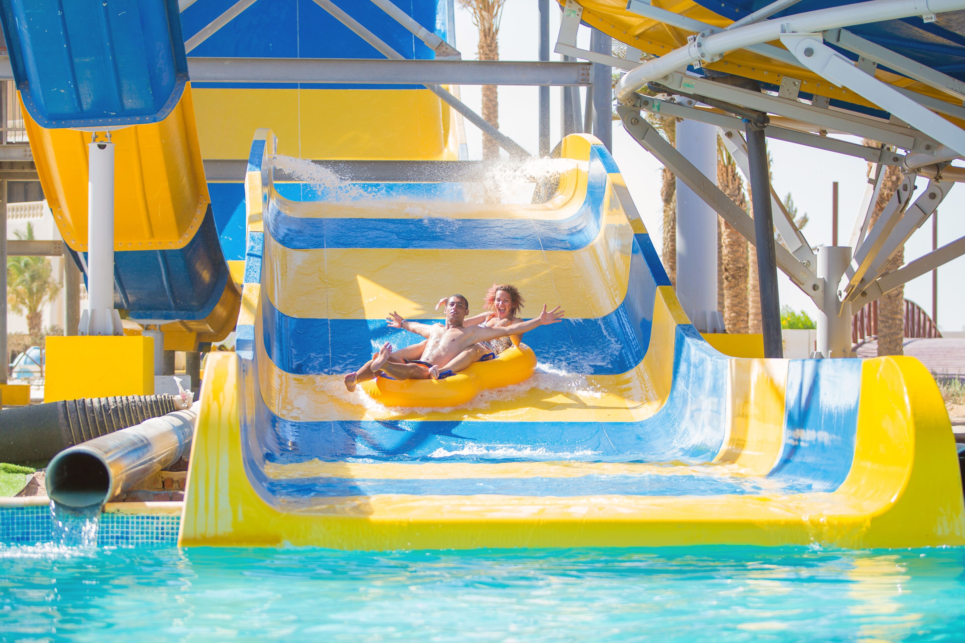 A water slide with two people at the bottom, a pool, and a structure with yellow and blue colors.
