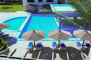 A pool area with multiple sun loungers and umbrellas, a separate smaller pool, and a view of a building in the background.