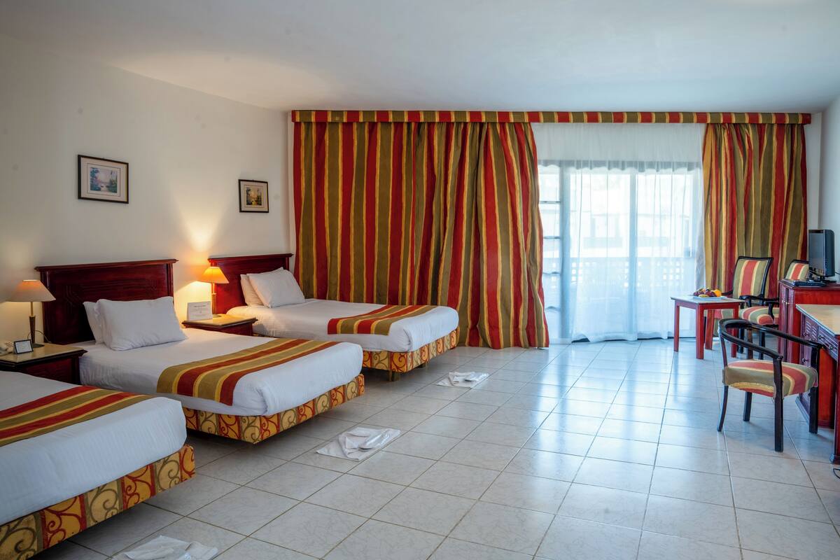 A hotel room with two beds, a desk with a chair, a television, and a large window with curtains.