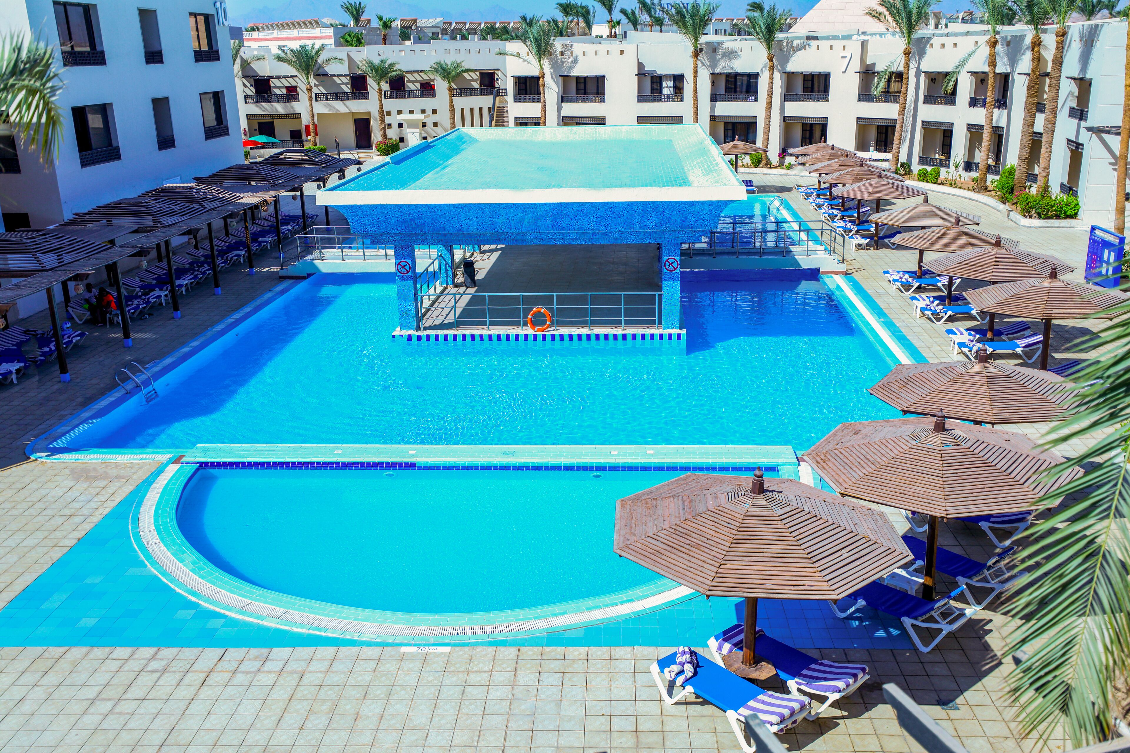 Ein Hotelpoolbereich mit mehreren Liegestühlen und Sonnenschirmen, einem zentralen Pool mit Sprungbrett und einer umlaufenden Poolterrasse.