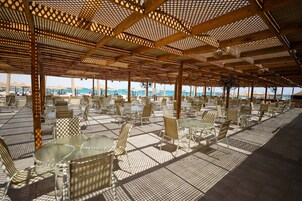 Un área de estar al aire libre con mesas y sillas, techo con diseño y vista a la playa y al océano.