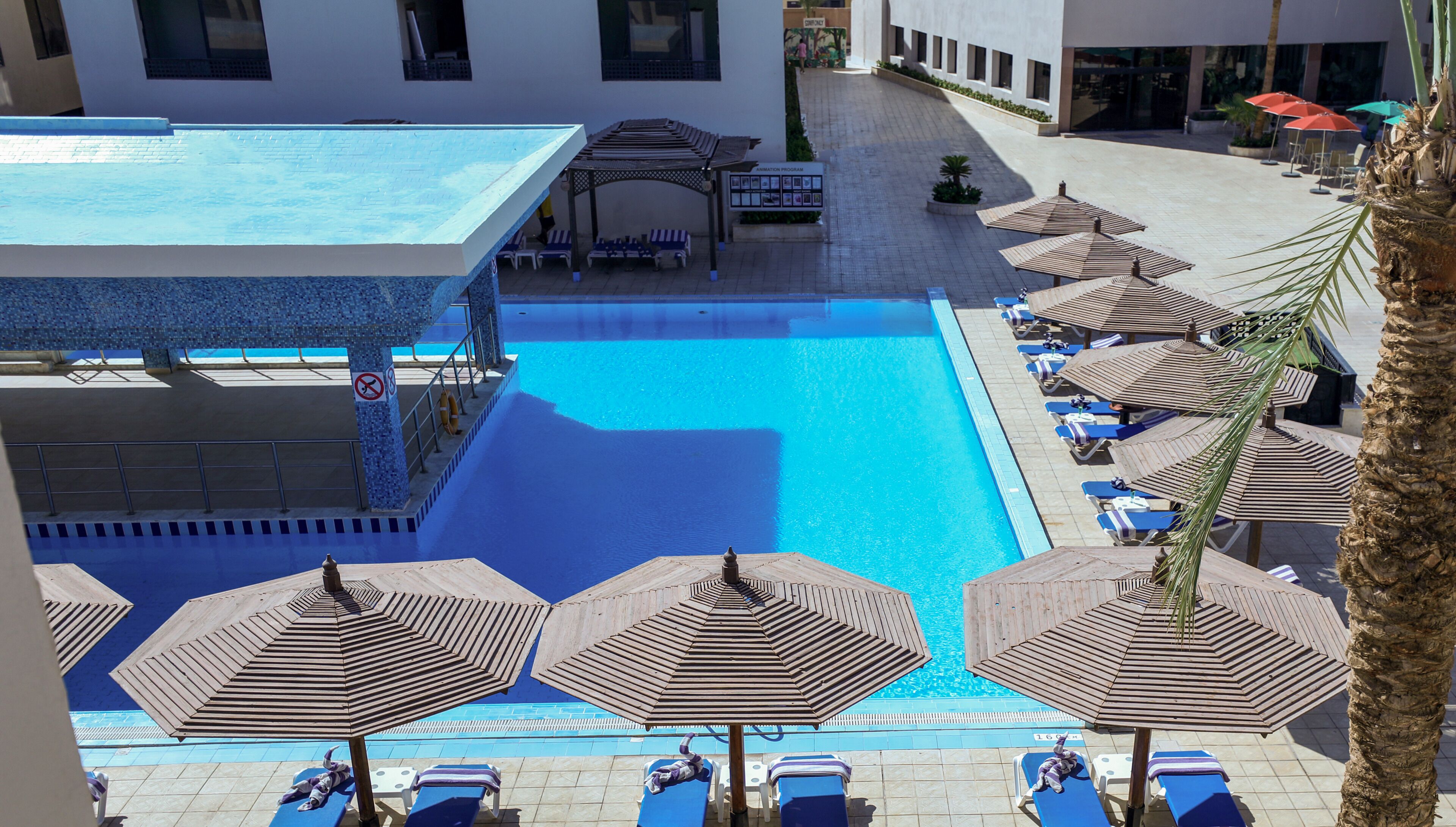 Uma área de piscina com espreguiçadeiras e guarda-sóis, um espaço coberto para sentar e uma placa indicando um 'Resort Program'.