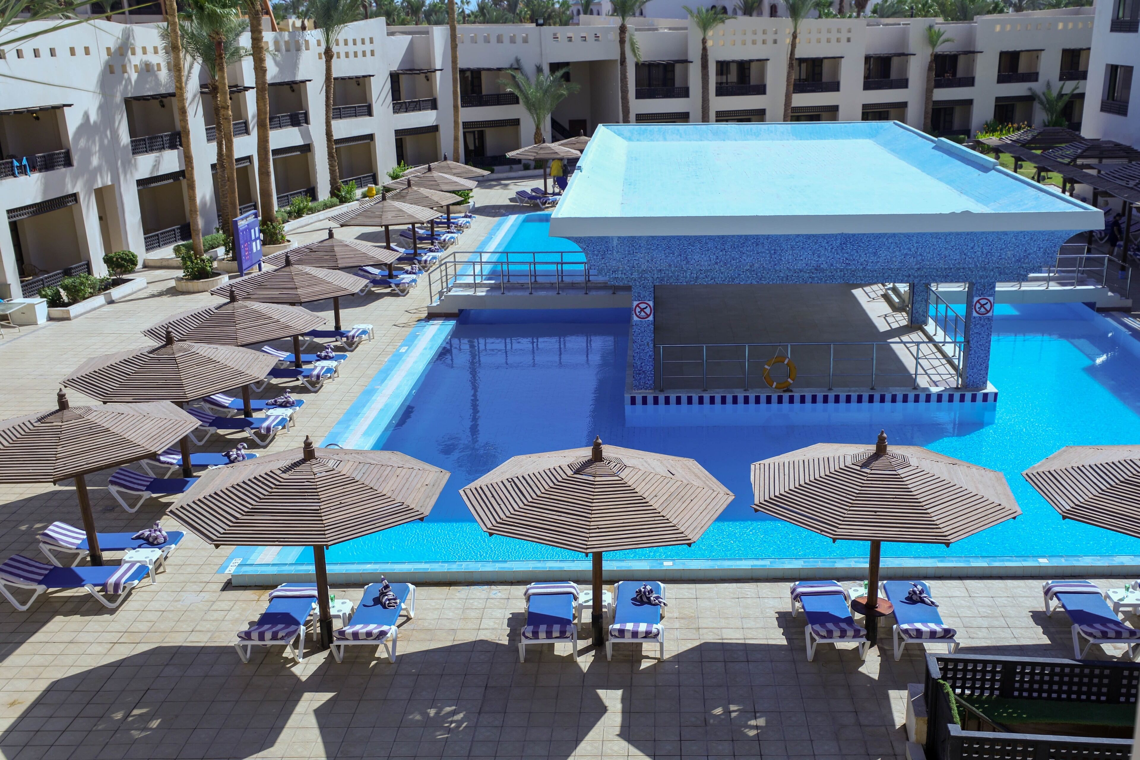 A hotel pool area with multiple lounge chairs and umbrellas, a central pool, and a surrounding building with balconies.