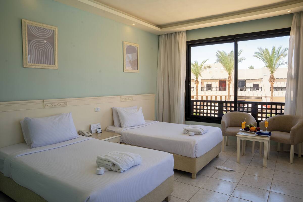 A hotel room with two beds, a small table with fruit and drinks, a view of palm trees and buildings outside, and a balcony with a lattice fence.