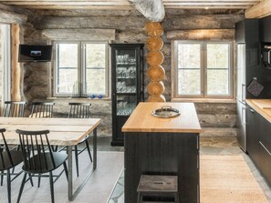 A rustic interior with a wooden dining table, chairs, a built-in kitchen island, and a glass cabinet with wine glasses.