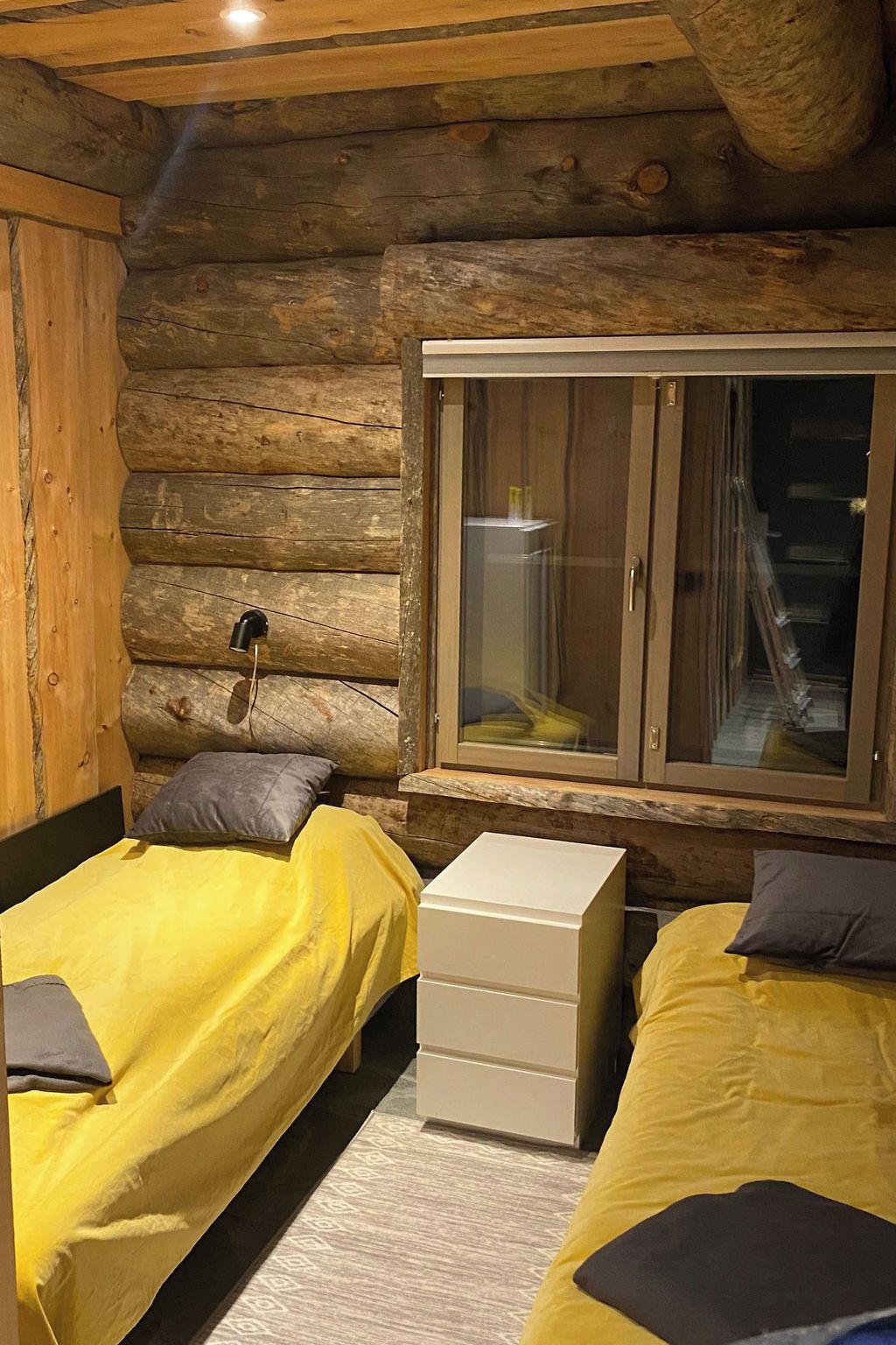 Een blokhutkamer met twee bedden, een klein wit dressoir en een raam met uitzicht op een trap.