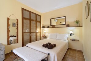 A neatly made bed with towels, a bedside table with a lamp, a mirror, and a wall-mounted shelf with a plant.