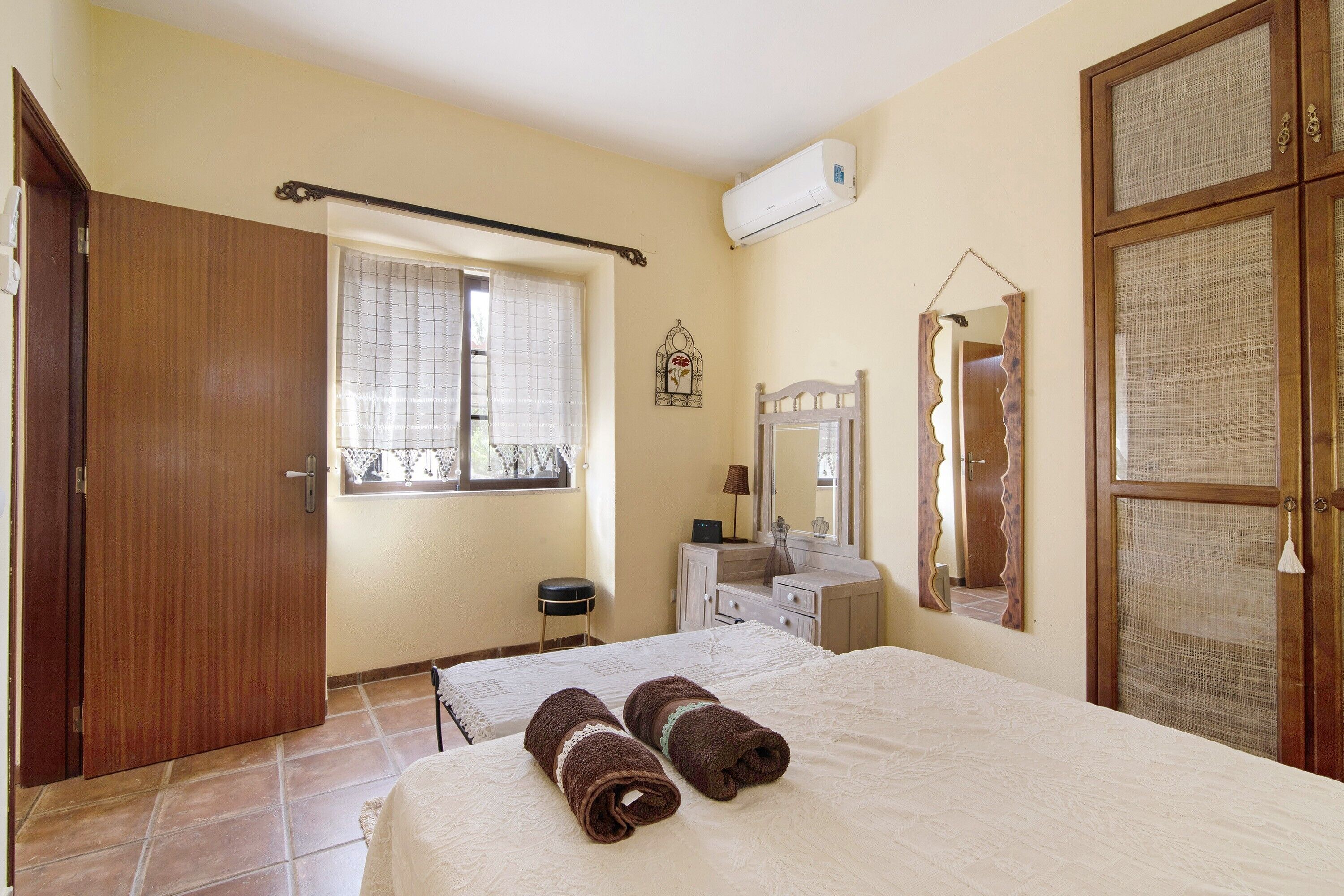A bedroom with a bed, a wooden door, a window with a curtain, a small stool, a vanity with a mirror, and an air conditioning unit.
