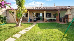 Une maison avec une façade en pierre, un toit en tuiles, un patio avec des meubles d’extérieur et un jardin agrémenté d’un palmier.