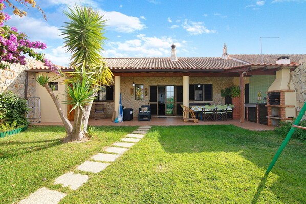 A house with a stone facade, a tiled roof, a patio with outdoor furniture, and a garden with a palm tree.