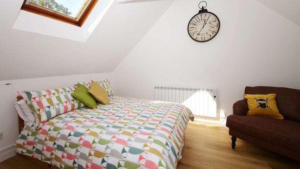 Een slaapkamer met een schuine kap, een bed met kleurrijk beddengoed, een bruine bank met een geel kussen en een wandklok.