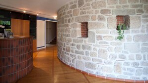 Un muro de piedra curvado con una pequeña ventana y una planta, suelo de madera y una zona de recepción con mostrador.