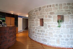 A curved stone wall with a small window and a plant, a wooden floor, and a reception area with a counter. - Logis Hotel de la Tour (Dunieres)