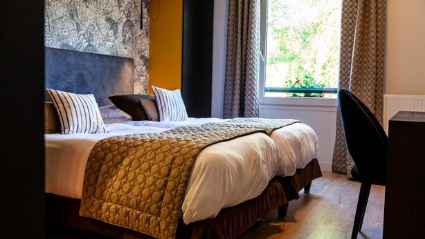 A neatly made bed with a patterned blanket, a headboard with a floral design, and a window with curtains. - Logis Hotel de la Tour (Dunieres)