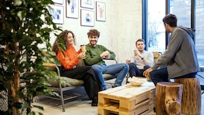 A group of people sitting on a couch in a modern, well-lit room with a large window, wooden furniture, and framed pictures on the wall. - Ten to Ten Hostel (Sapporo)