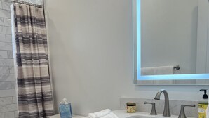 Un baño moderno con un inodoro blanco, una ducha con cortina a rayas, un lavamanos blanco con espejo encima y un mueble de tocador con cajones y un perchero.