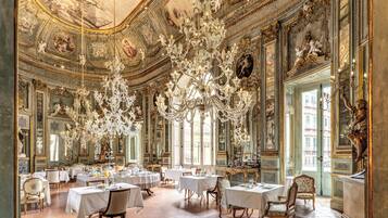 An opulent dining room with ornate chandeliers, multiple dining tables, and classical artwork on the walls.
