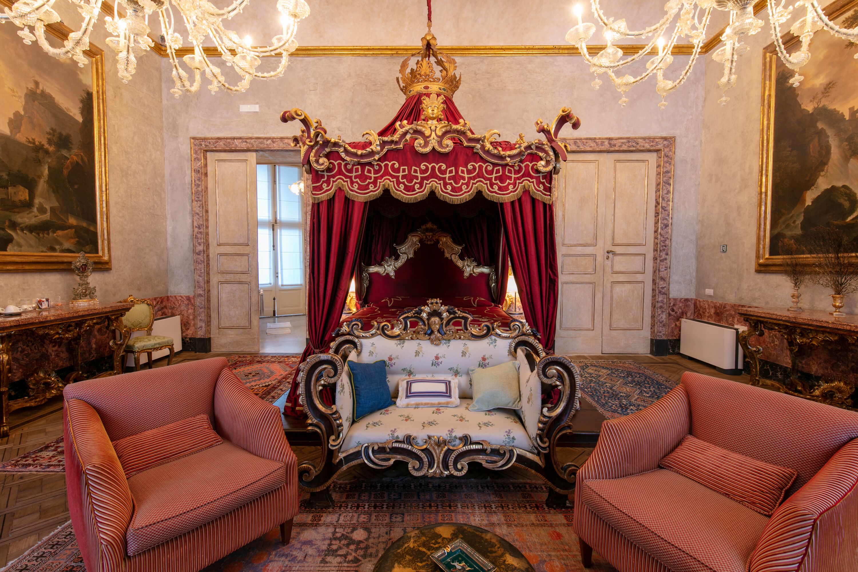 A luxurious bedroom with a four-poster bed, ornate curtains, and a chandelier. There are two upholstered chairs and a large painting on the wall.