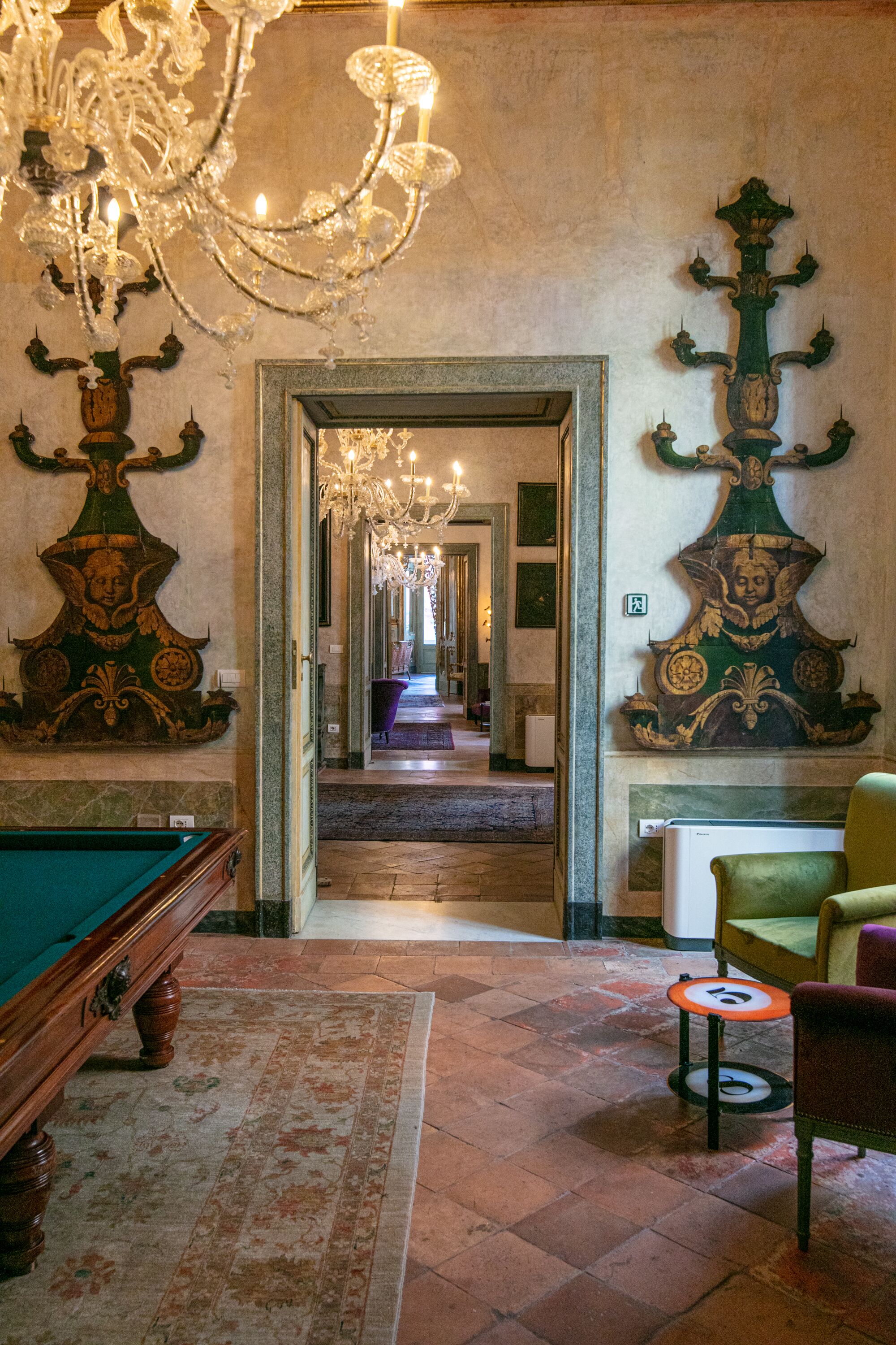 Una mesa de billar, un sillón verde y una araña en una habitación con decoraciones elaboradas en las paredes y un pasillo que lleva a otra estancia.