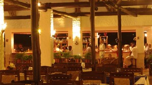 An indoor restaurant with wooden tables and chairs, a bar area with patrons, and potted plants. - Mnarani Beach Club (Malindi)