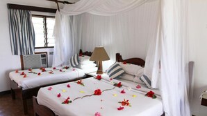 A hotel room with two beds, each with white linens and floral decorations. There is a bedside table with a lamp, a window with curtains, and an air conditioning unit. - Mnarani Beach Club (Malindi)