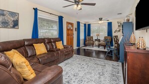 A living room with a brown leather sofa, blue curtains, a ceiling fan, and a patterned rug.