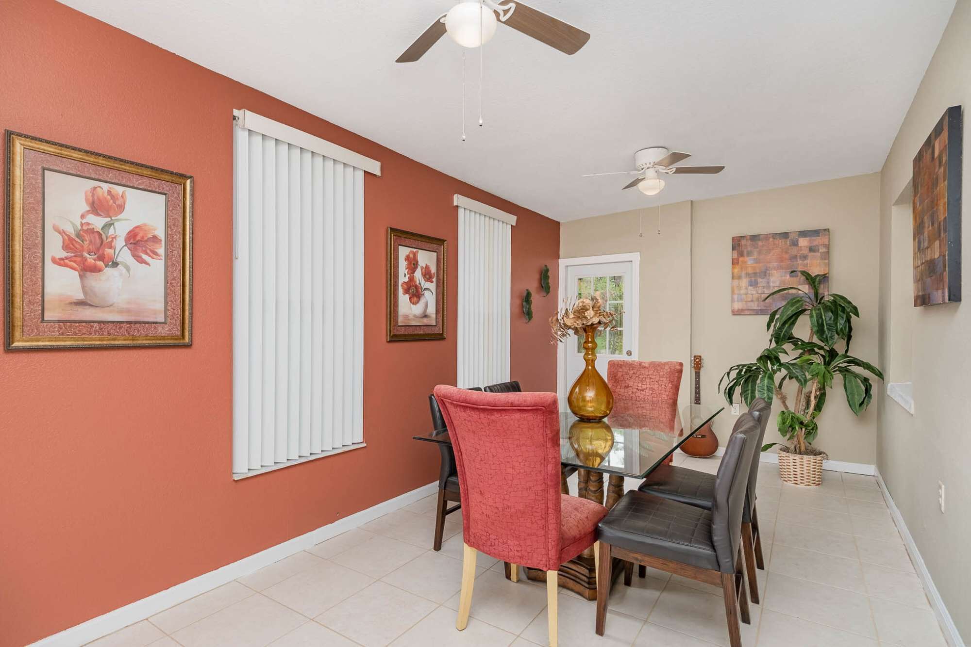 A dining area with a glass table, red and black chairs, ceiling fans, and a potted plant.