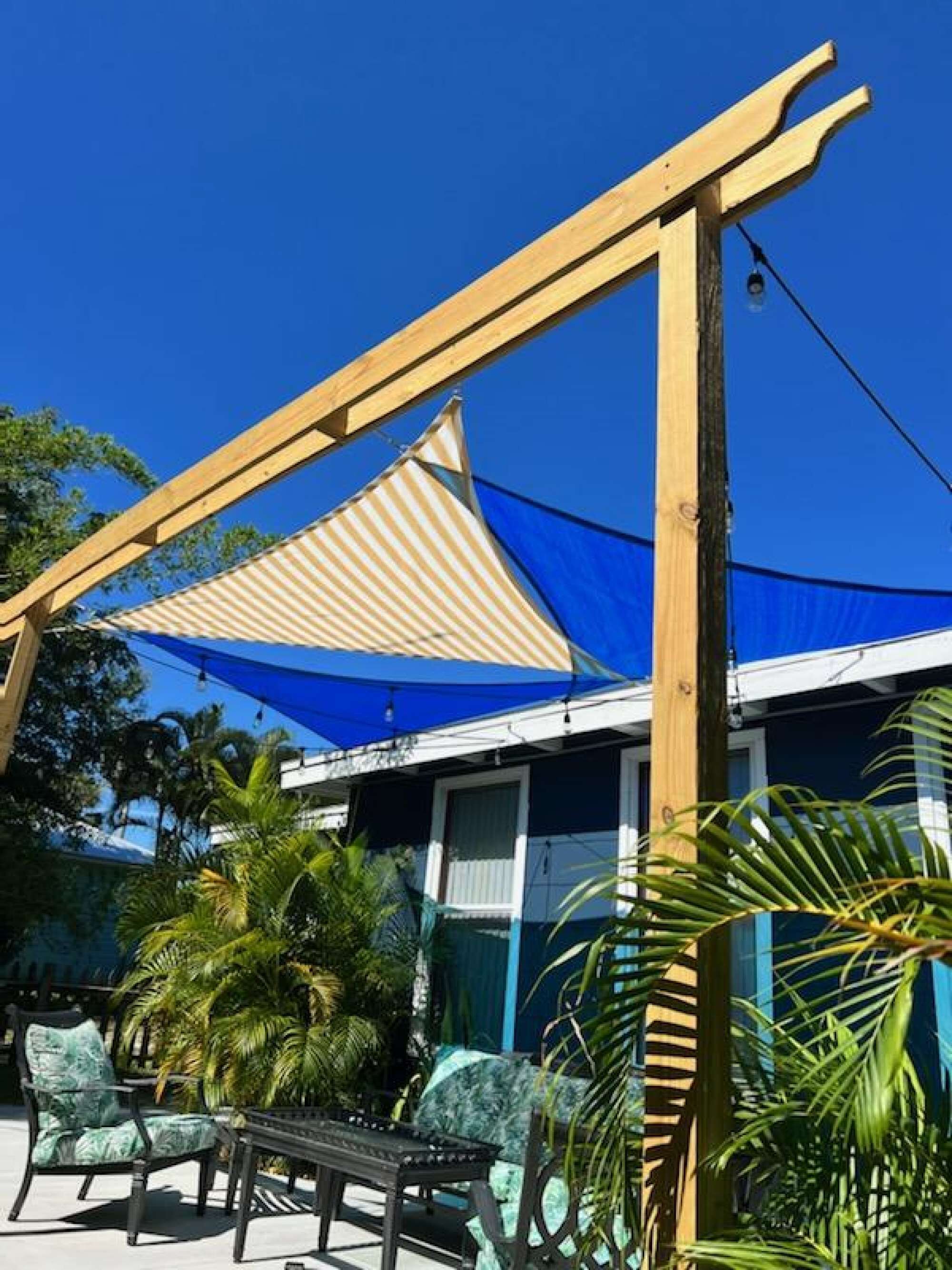 Una pérgola de madera con toldos de tela a rayas, mobiliario exterior y plantas tropicales.