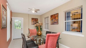 A dining area with a glass table, chairs, a ceiling fan, and artwork on the walls.