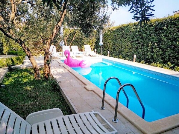 Relax by the pool surrounded by lush greenery and a shaded tree.