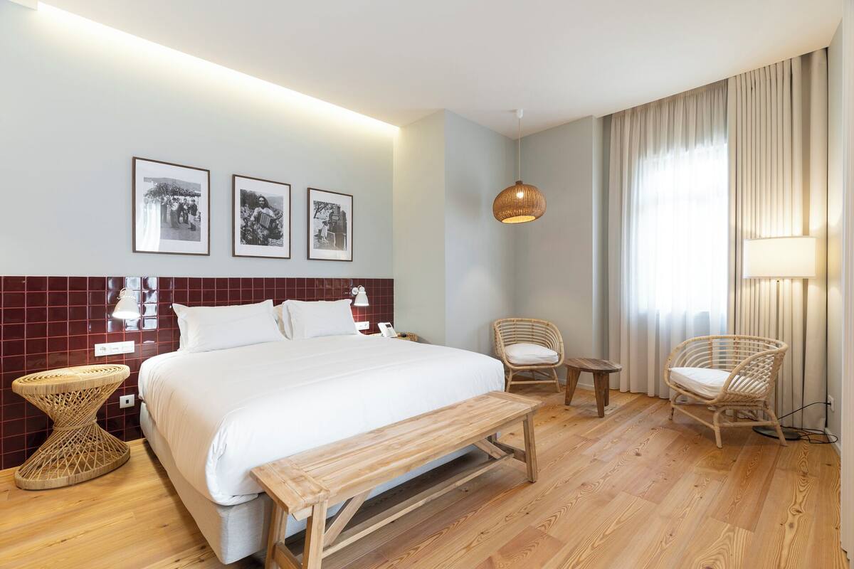 Ein Hotelzimmer mit einem großen Bett, einer hölzernen Bank, zwei Korbstühlen und einem kleinen Tisch, sowie einer Wandleuchte.